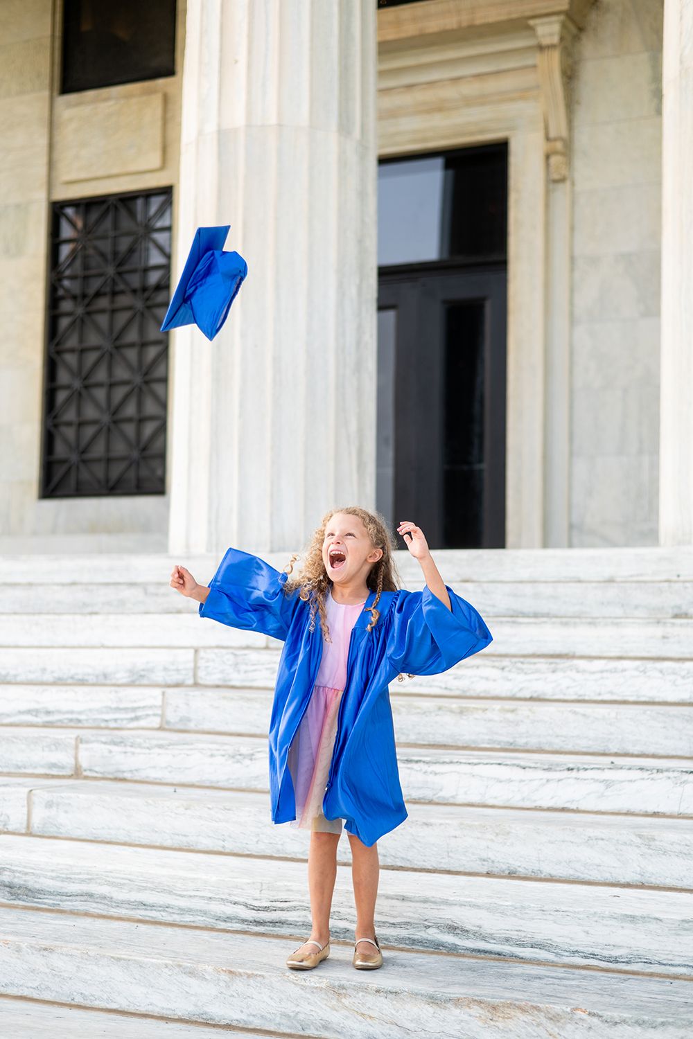 Graduate celebrates by tossing blue cap in the air while standing on white marble steps.
