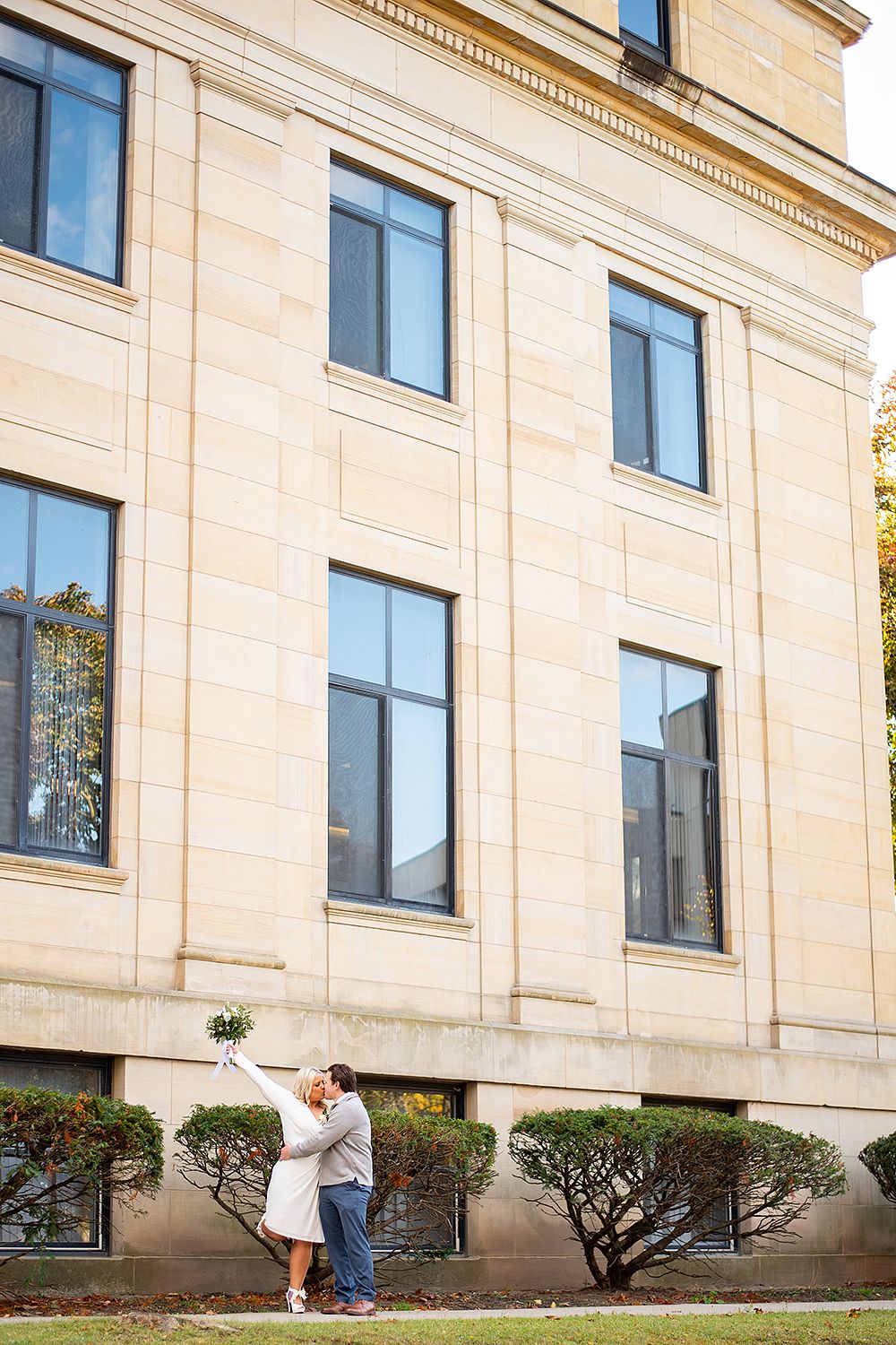 Couple embraces in front of a beige stone building with tall windows and manicured shrubs along the foundation.