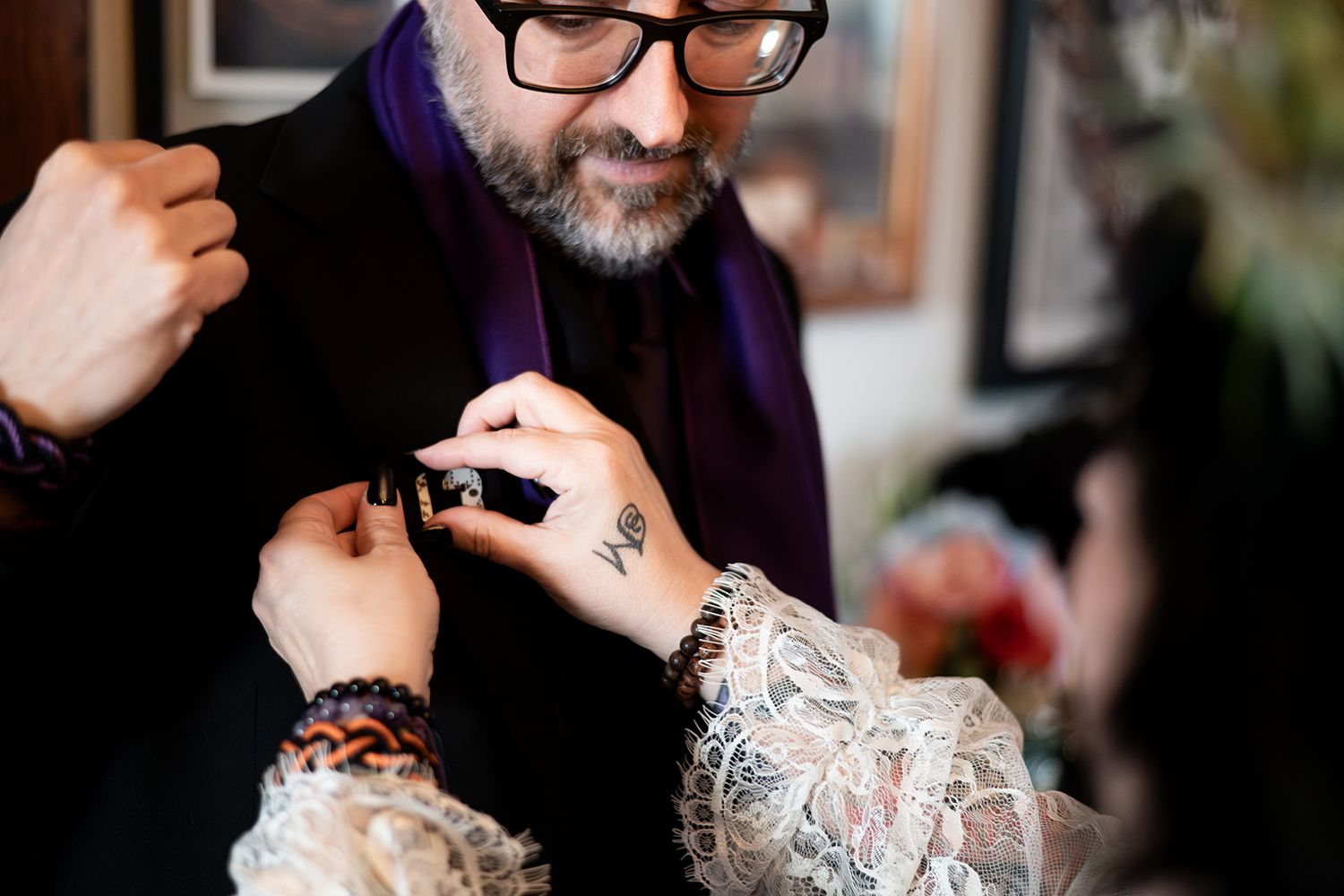 A tattoo artist wearing glasses and a purple scarf works meticulously on a client's detailed tattoo artwork.