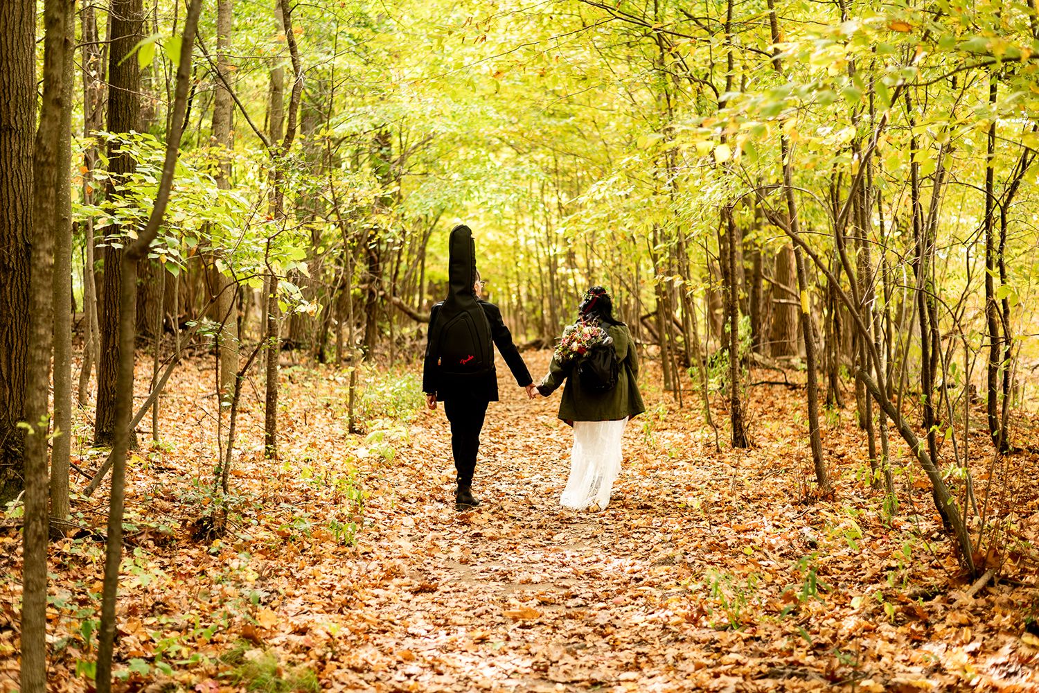 Two people walk along a leaf-covered trail through golden autumn woods on a sunny afternoon.