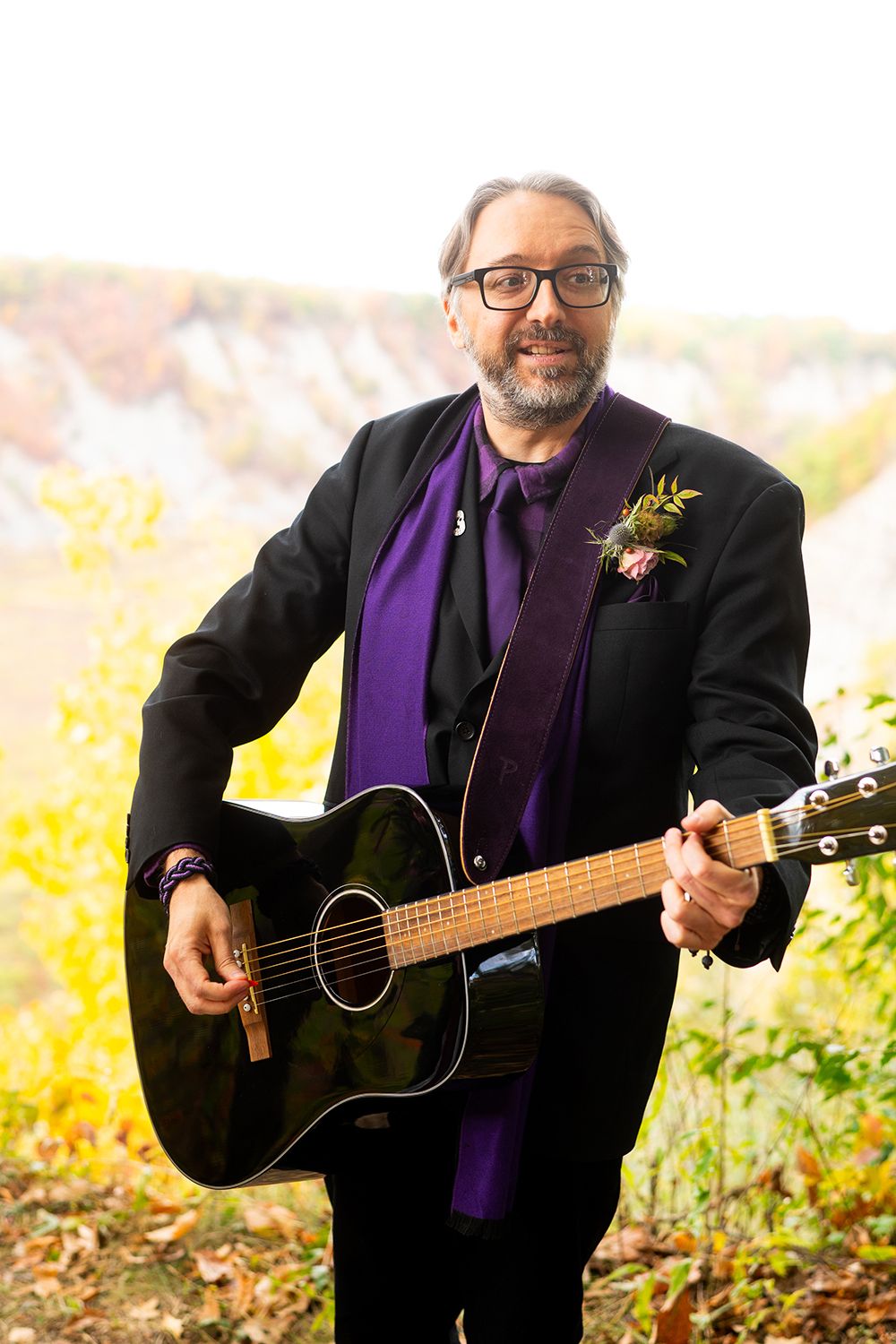 A musician in a black suit and purple tie holds an acoustic guitar outdoors against a scenic autumn backdrop.