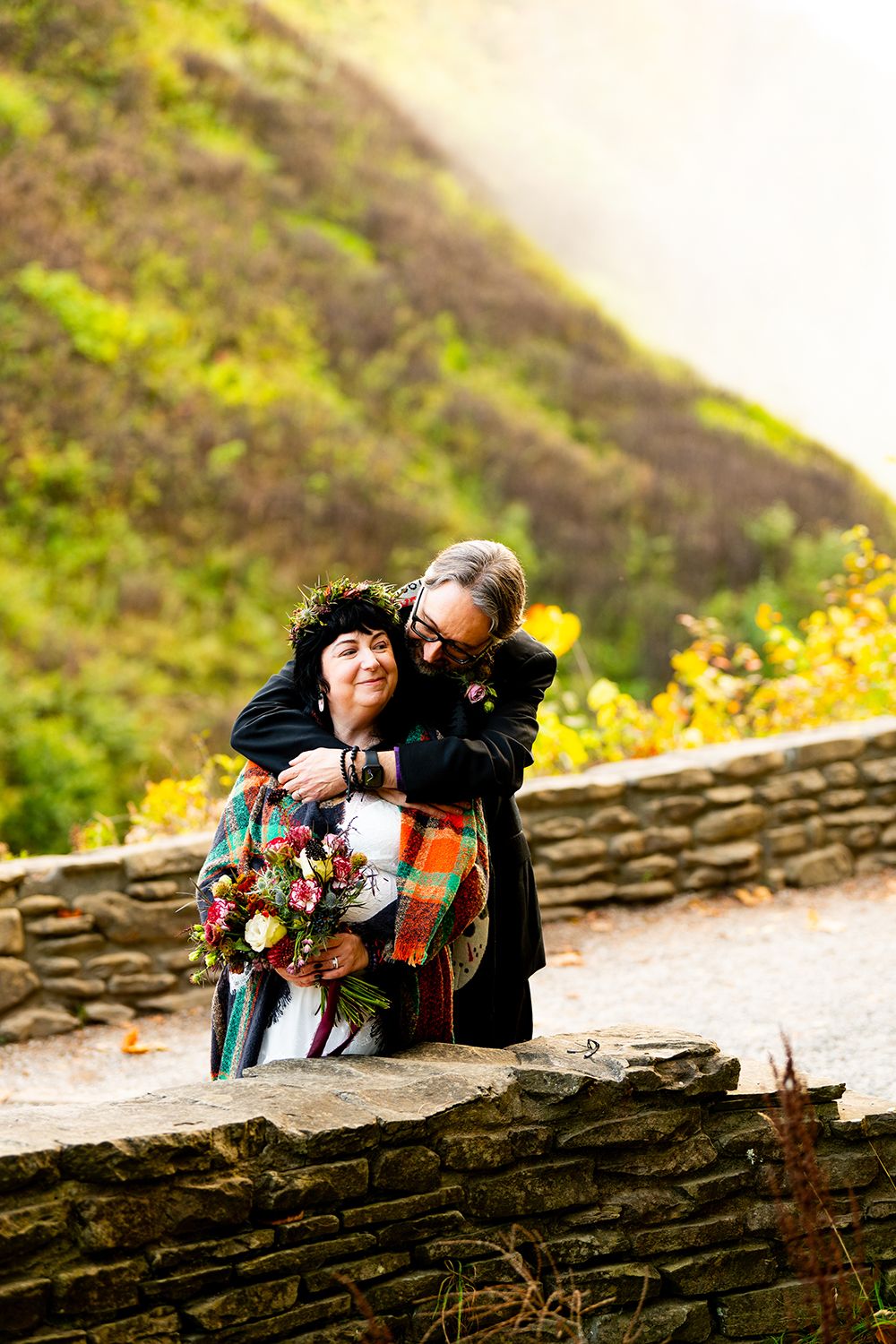 A loving couple embraces while standing by a stone wall on a mountainside with vibrant yellow wildflowers in the background.