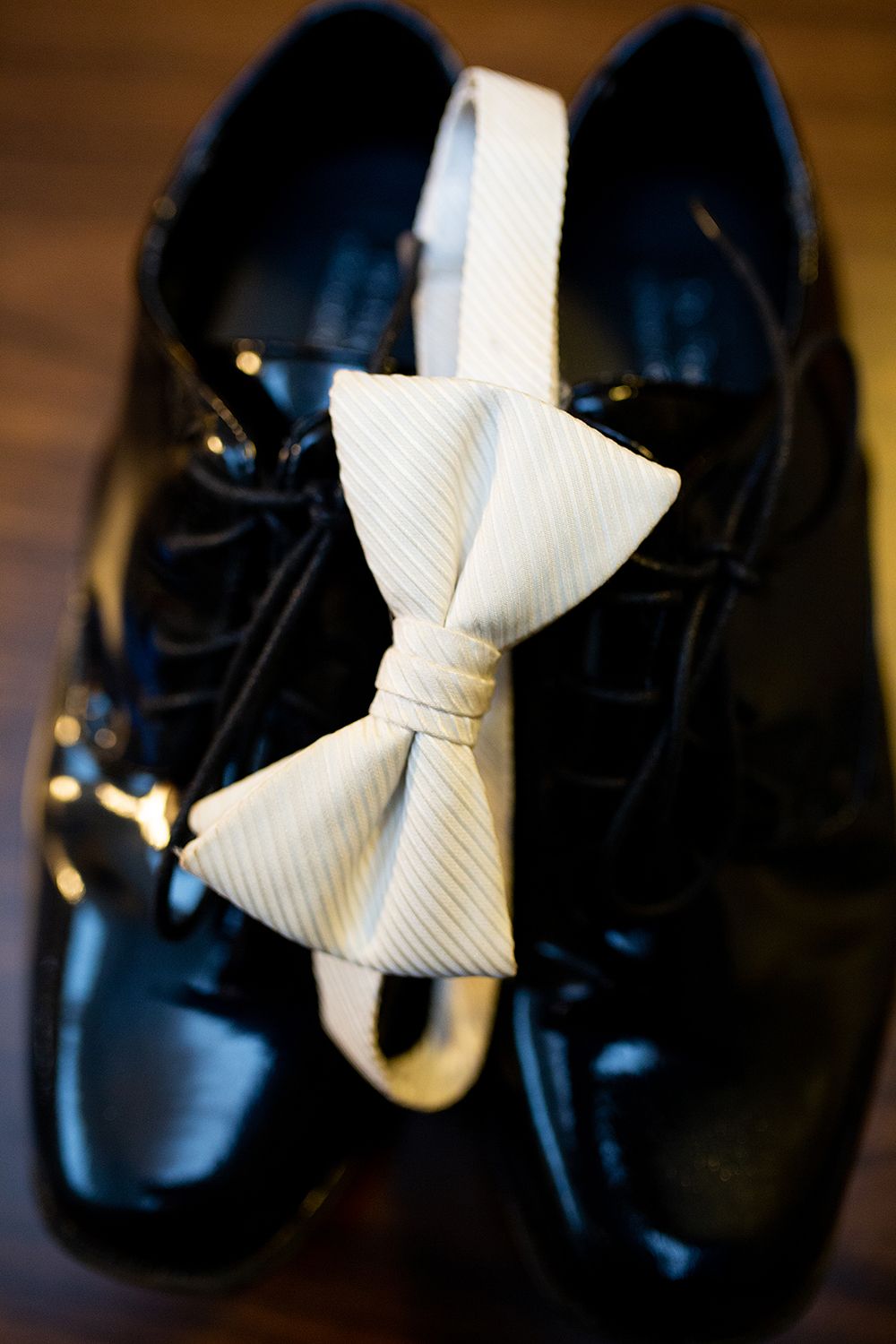 A white bowtie rests atop shiny black formal dress shoes on a dark wooden surface.