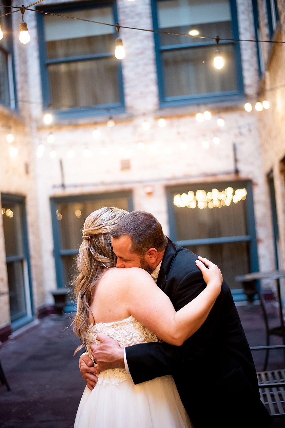 A couple in formal wedding attire share an intimate outdoor dance under string lights.