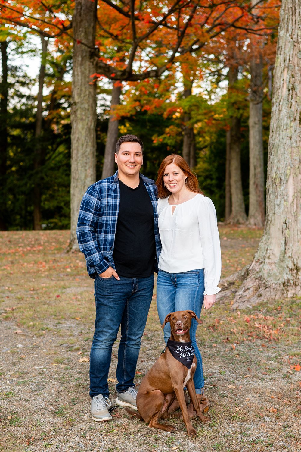 A couple poses with their brown dog in a fall forest setting with orange and yellow foliage.