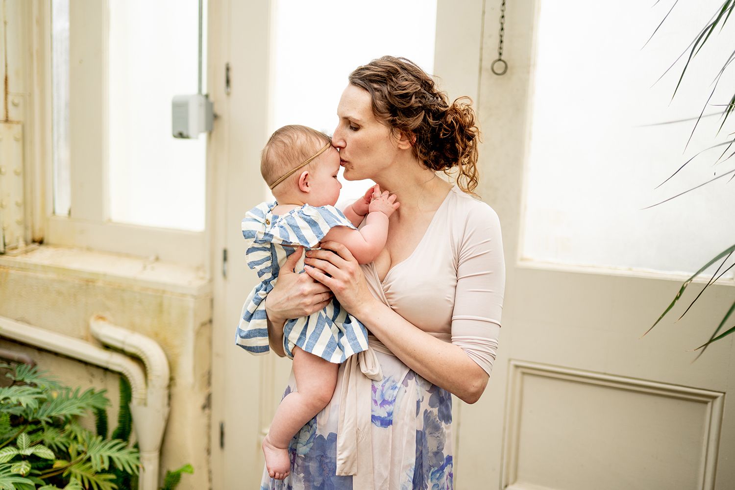 A parent in a floral dress lovingly holds and kisses a baby wearing blue striped clothing in a sunlit room.