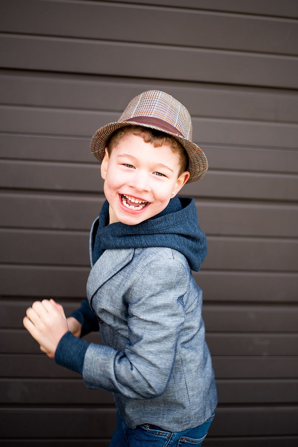 A joyful expression lights up a young subject wearing a plaid fedora and denim jacket against a gray wall.