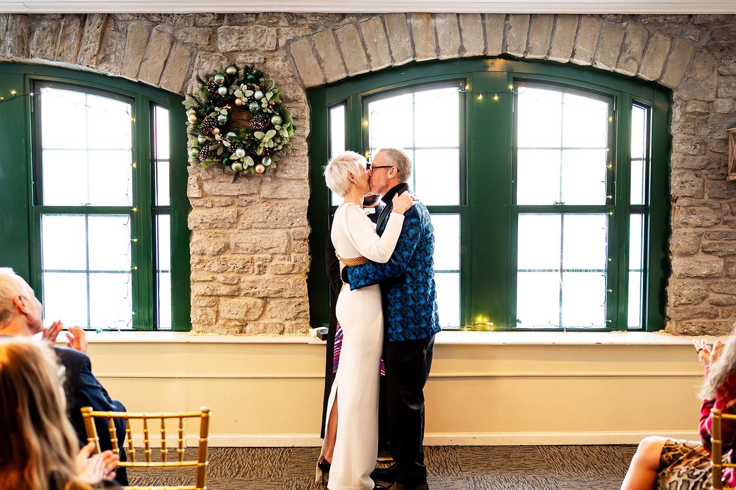 A couple embraces during an indoor ceremony with stone walls, green-trimmed windows, and a festive wreath decoration.