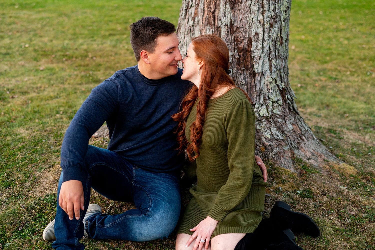 A couple in casual fall attire shares an intimate moment sitting together at the base of a tree.