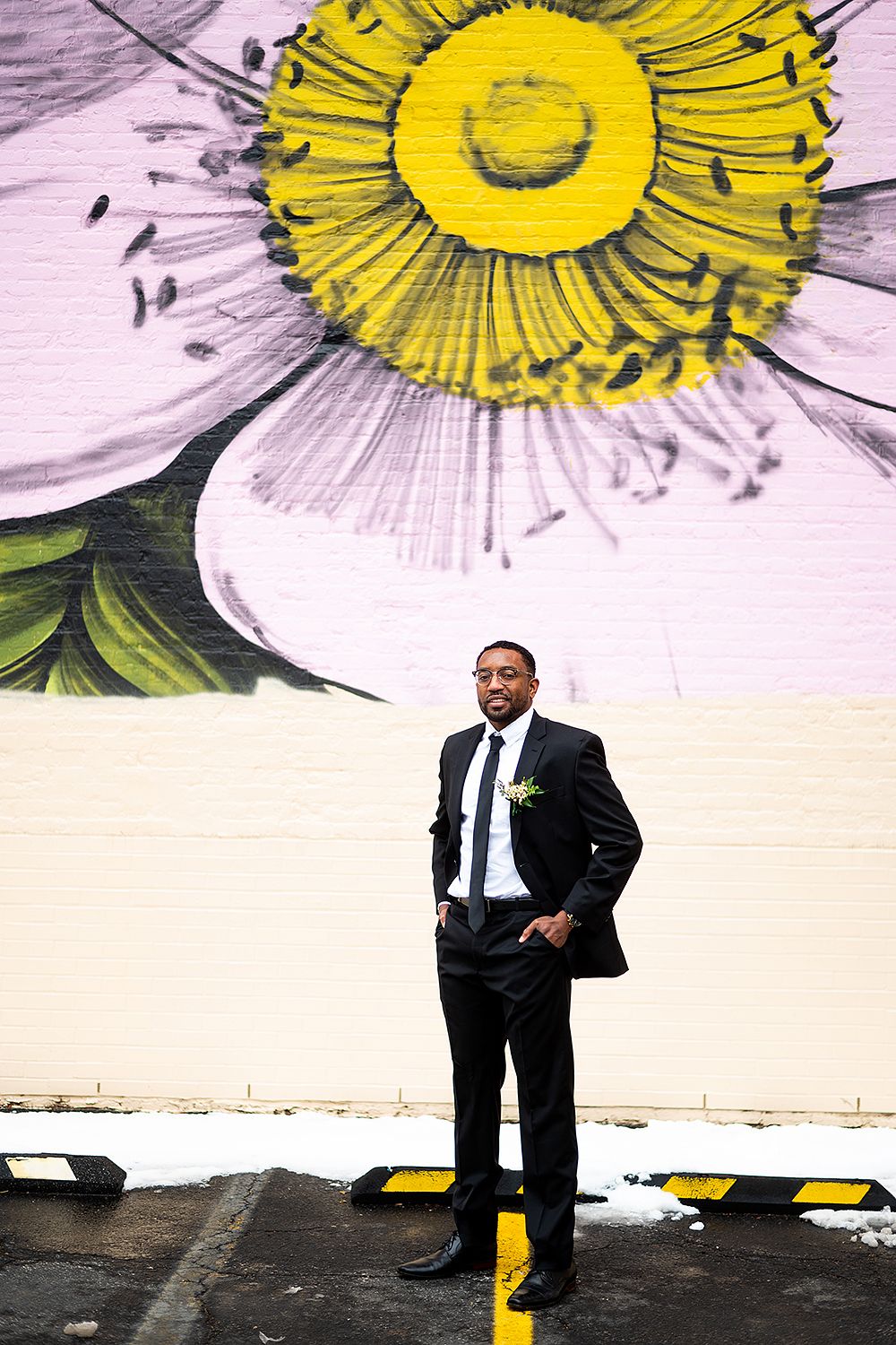 A well-dressed figure poses confidently against a wall with large painted white and yellow floral artwork.