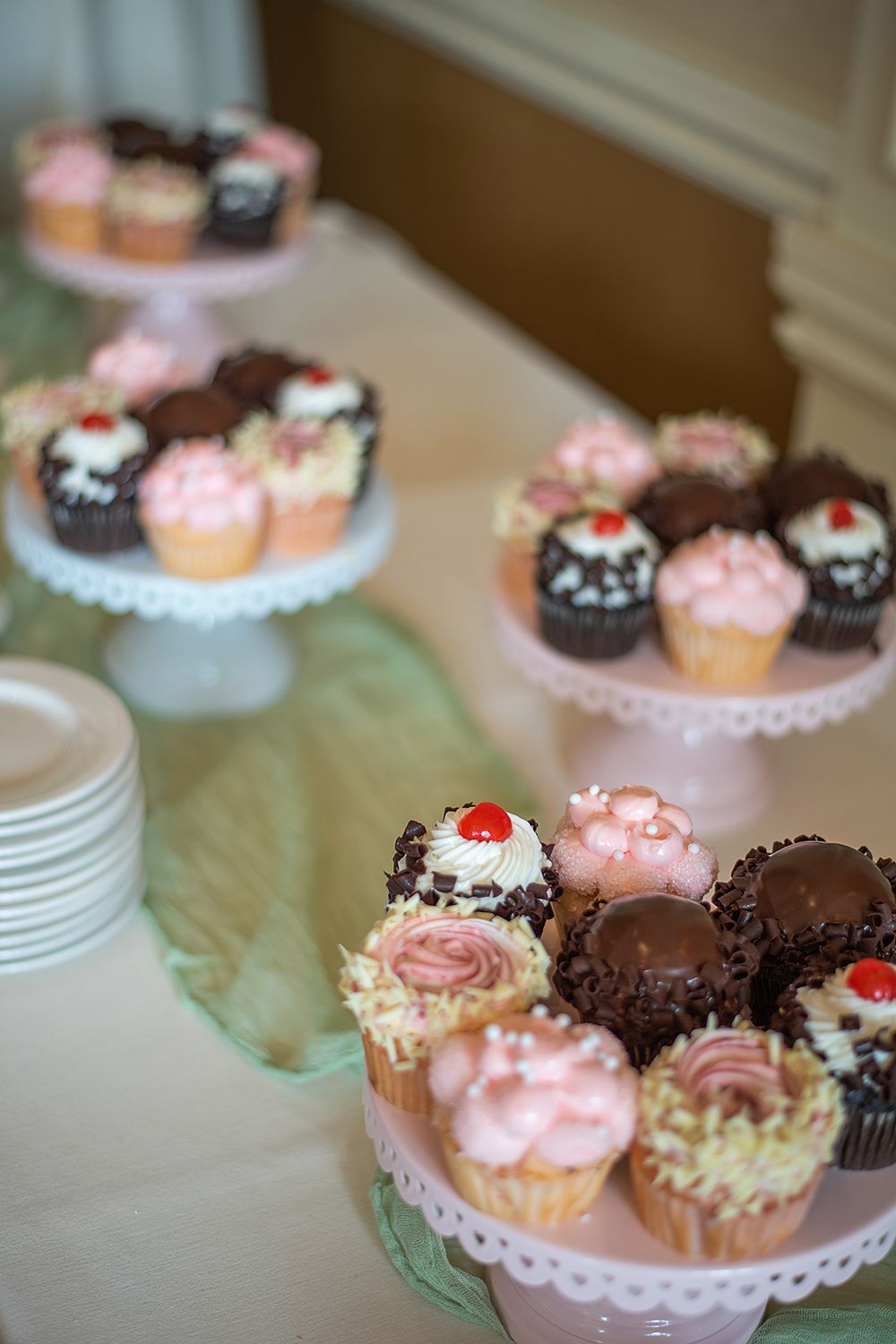 Assorted chocolate and pink frosted cupcakes displayed on tiered white lace cake stands at a dessert table.