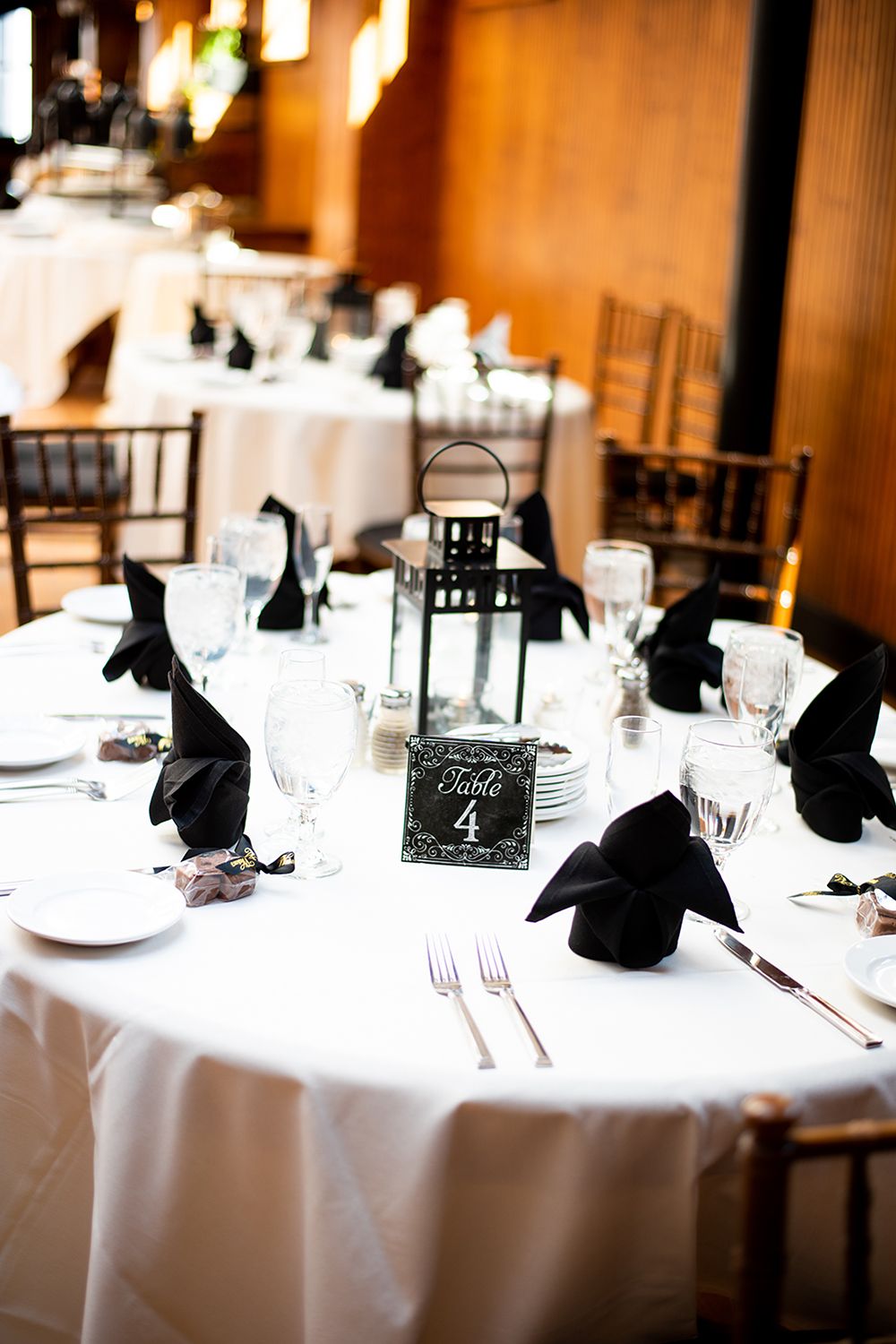 Elegant wedding reception table settings with white linens, black napkins and place cards in warm lighting.