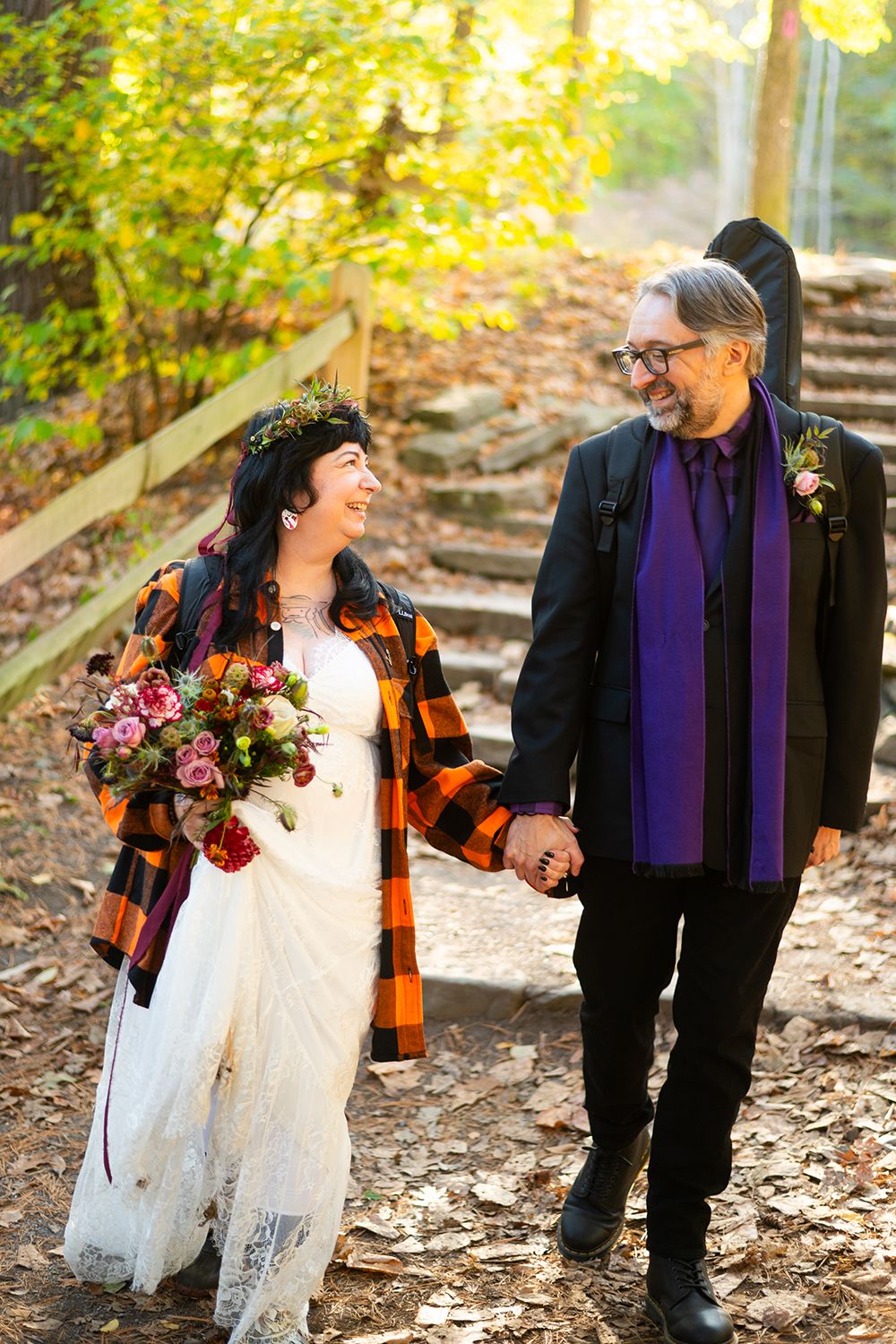 Couple in autumn outdoor wedding attire walking hand in hand on leaf-covered path with rustic plaid and boho style elements.