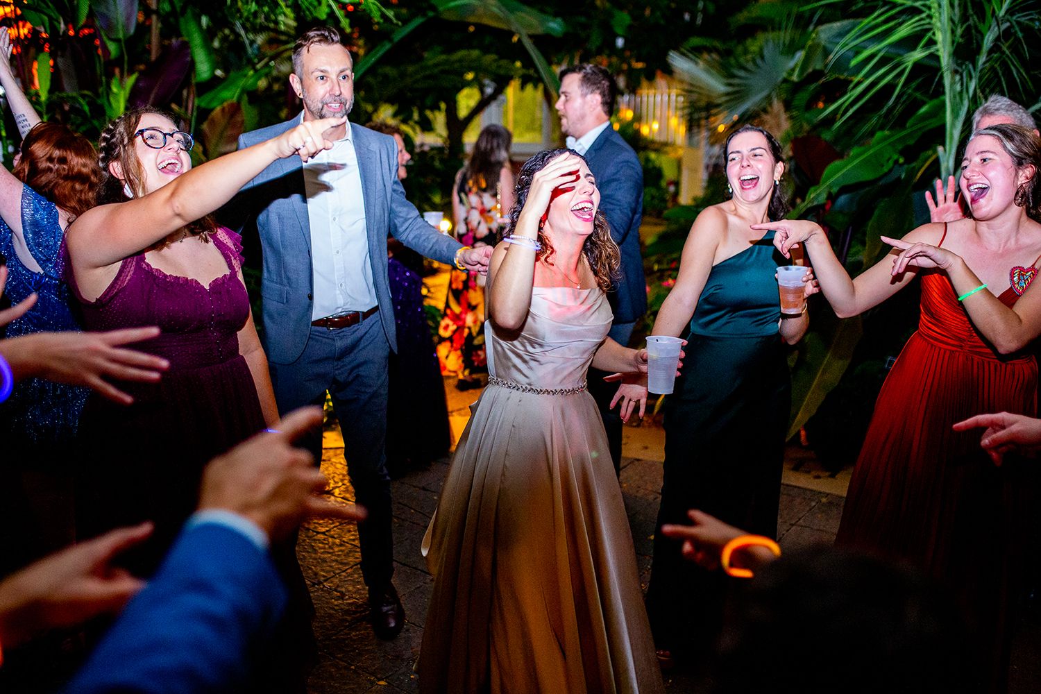 Wedding guests dance together on a festive dance floor surrounded by tropical plants at night.
