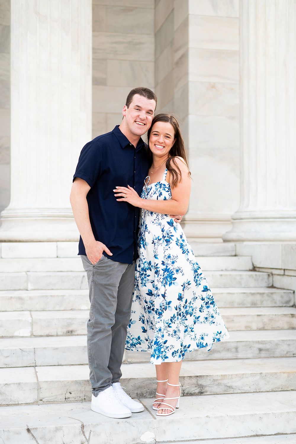 A couple poses on white marble steps, with one person wearing a blue floral dress and the other in a navy shirt.