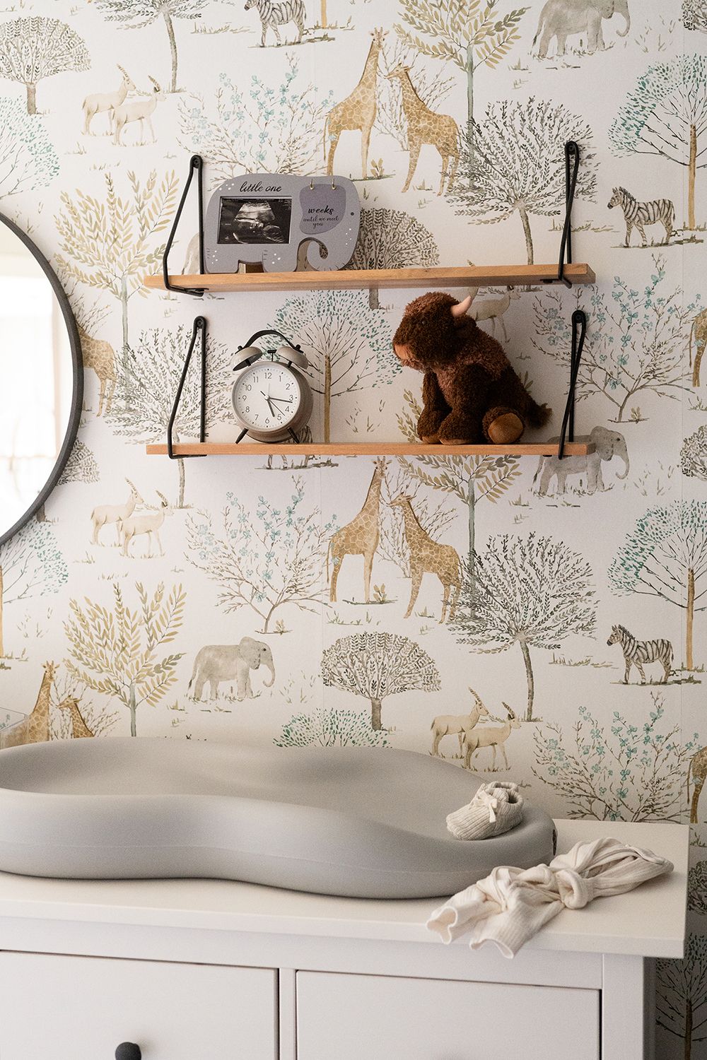 Nursery decor with wooden shelves, plush toy, and safari animal wallpaper featuring giraffes and elephants in muted tones.