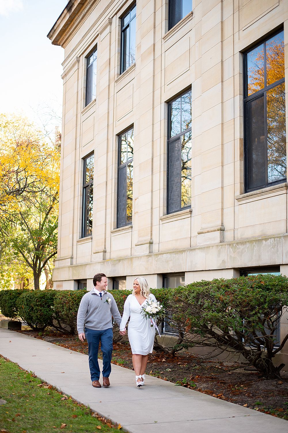 Couple walking outside a stone building with tall windows and autumn trees during engagement session.