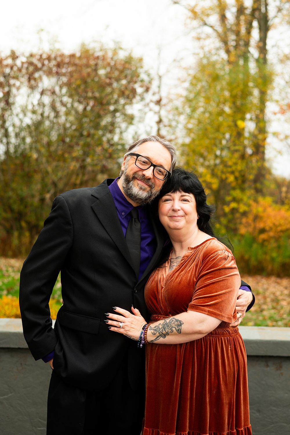 A couple embraces outdoors during autumn, wearing formal attire with a black suit and burnt orange velvet dress.