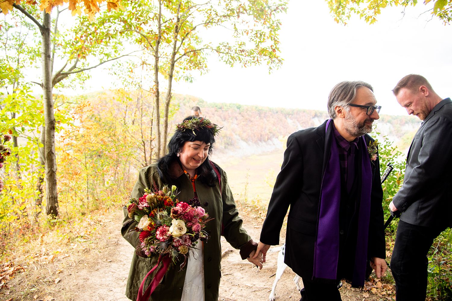 A couple walks hand in hand after their autumn wedding ceremony with fall colors in the background.