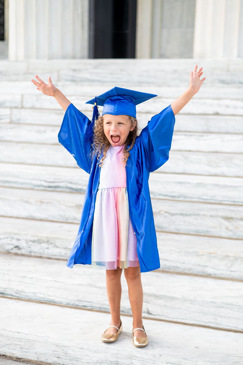 Small child in bright blue graduation cap and gown jumps excitedly on white marble steps.