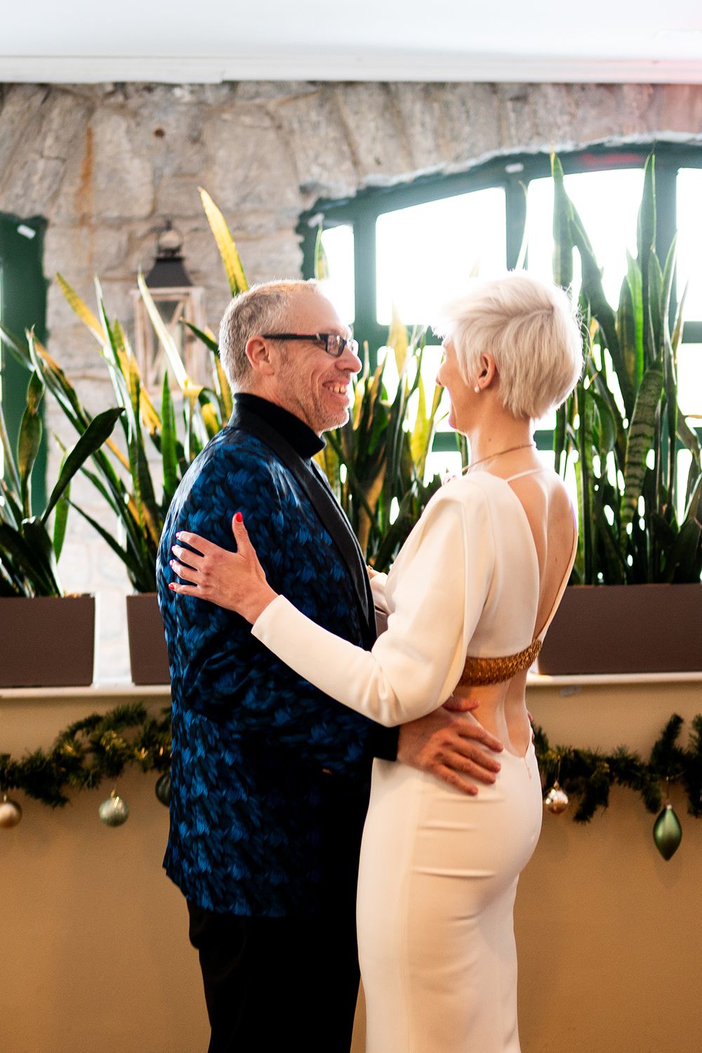 An elderly couple dances together in an indoor venue decorated with plants along a window ledge.