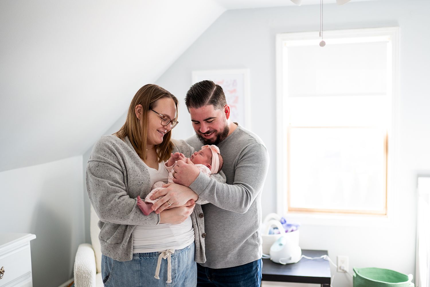 Two adults in casual sweaters share a tender moment holding a newborn baby in a bright white room.