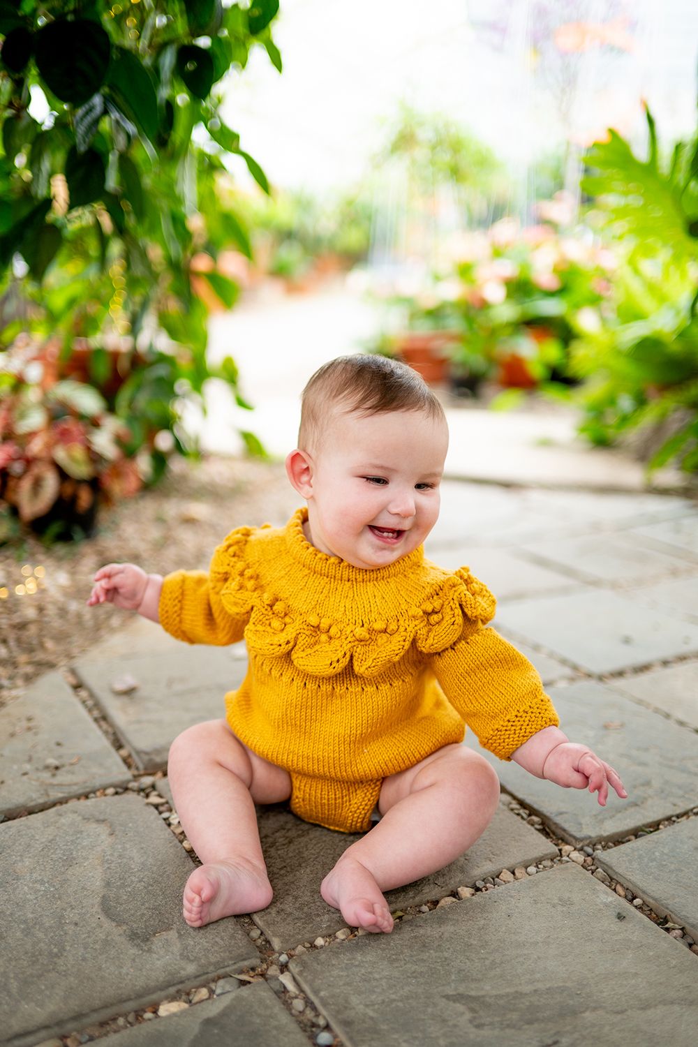 A happy baby wearing a mustard yellow knit sweater with scalloped detail sits on stone pavement in a garden setting.