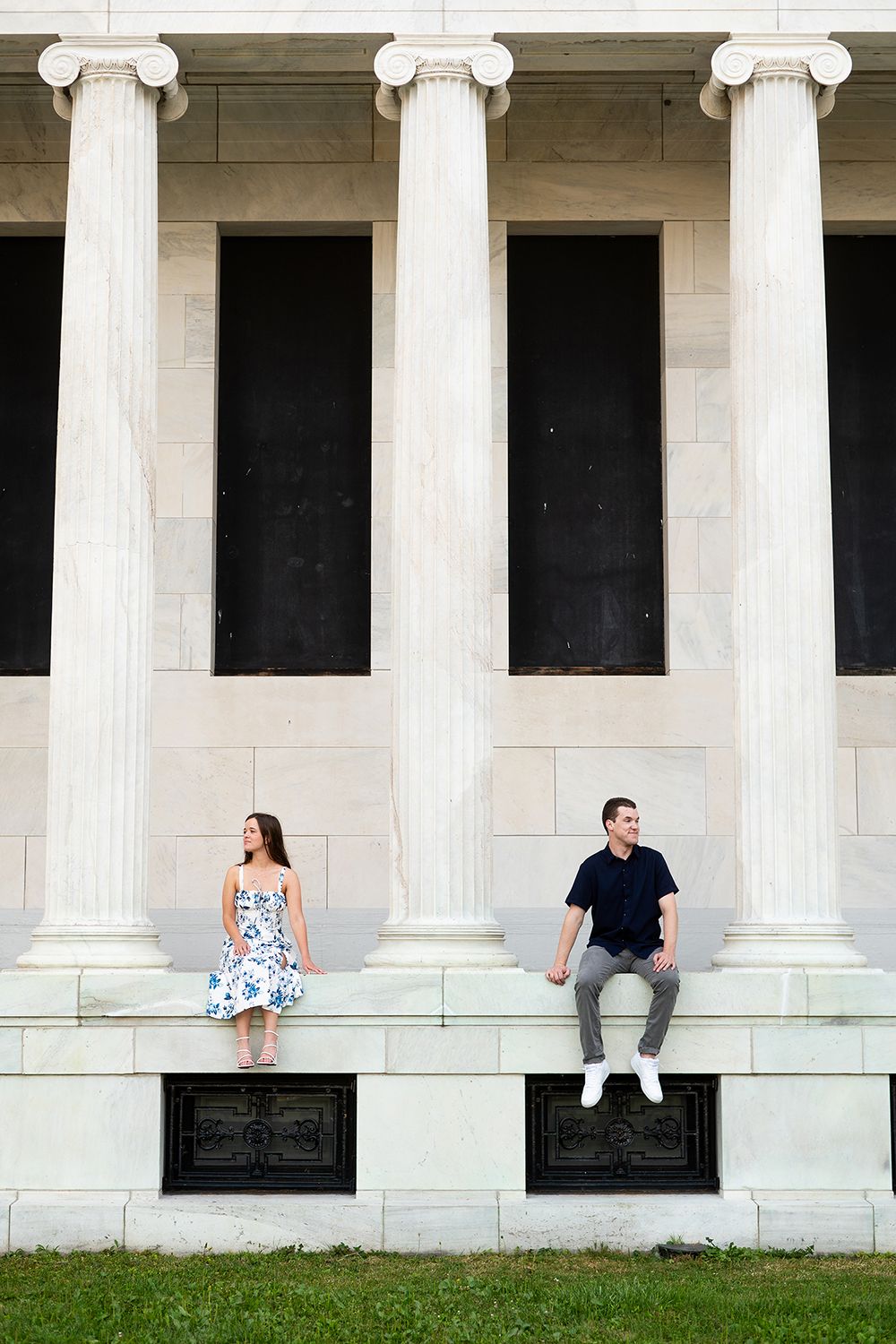 Two people sit apart on marble steps in front of classical white columns at a historic building.