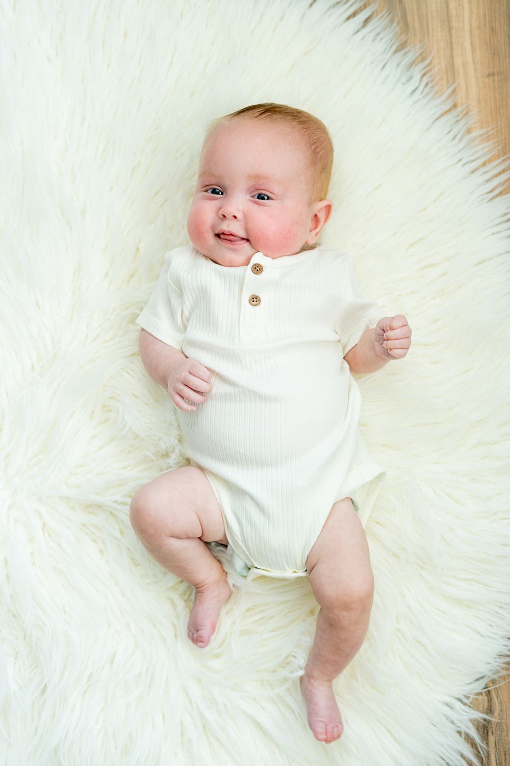 Baby wearing white ribbed onesie lies on soft white fur rug during portrait session.