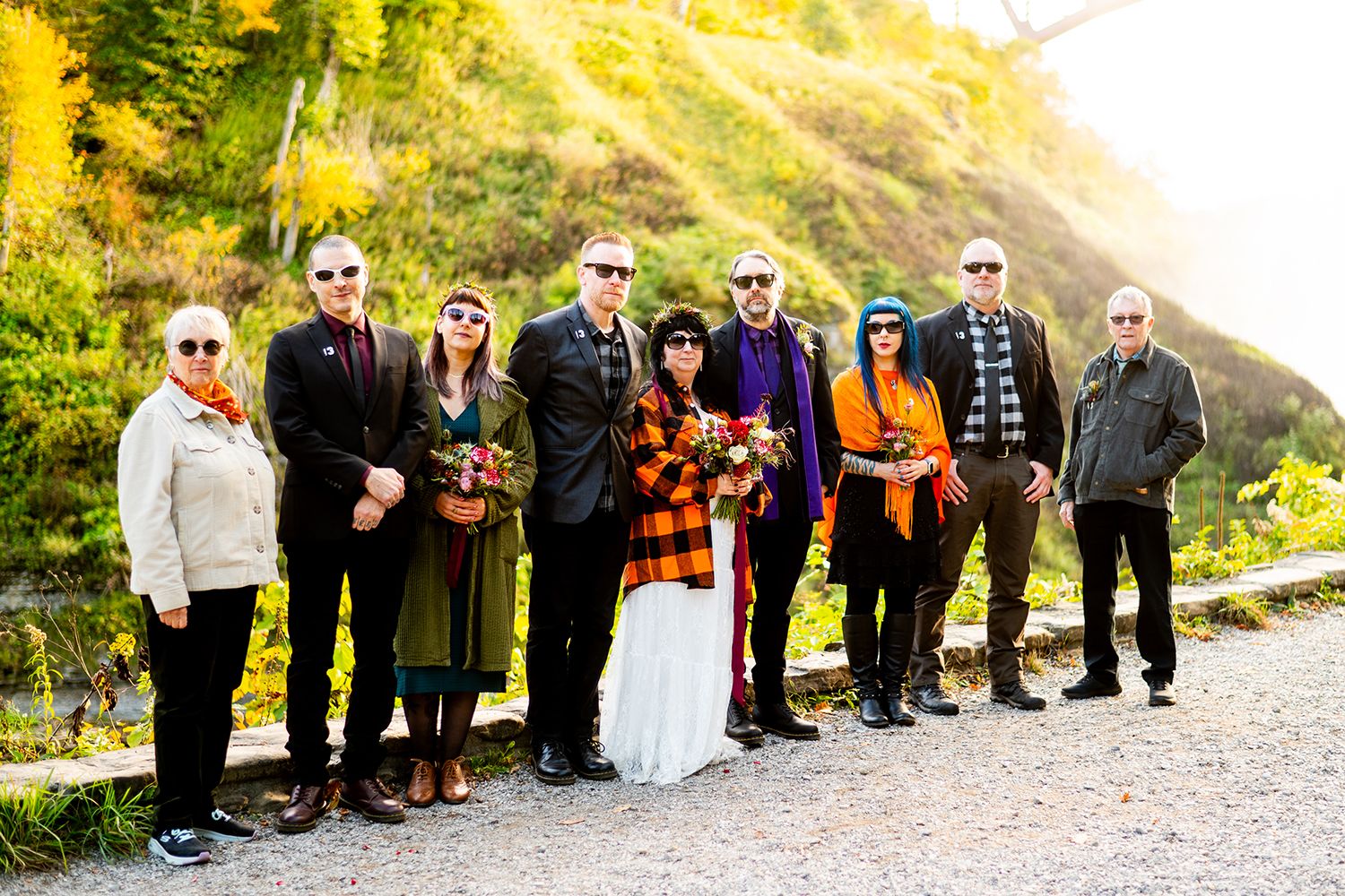 A lineup of wedding party members in black, orange and white attire poses against a green hillside at sunset.