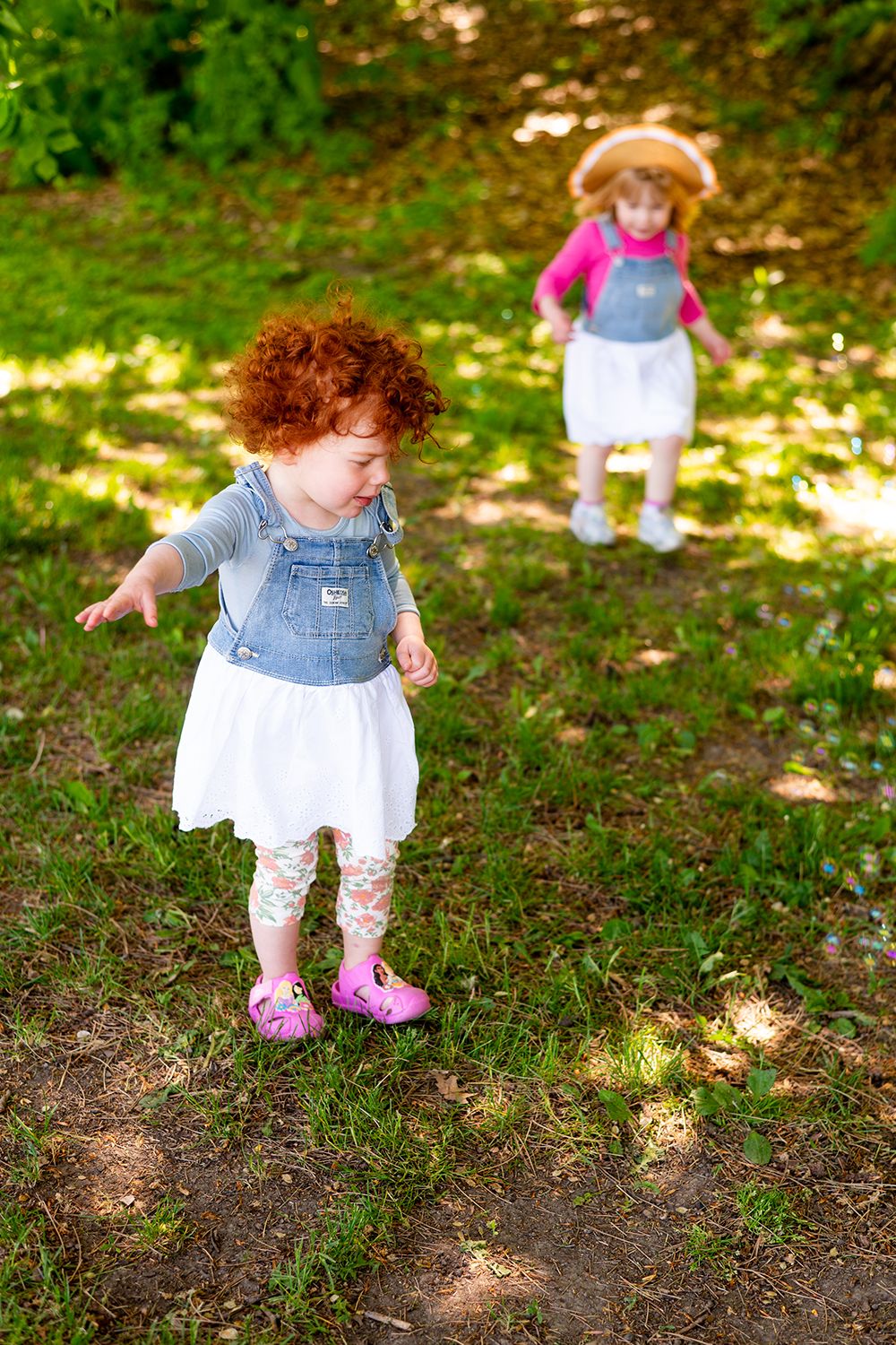 Two toddlers in denim overalls and dresses playing in a shaded wooded area.