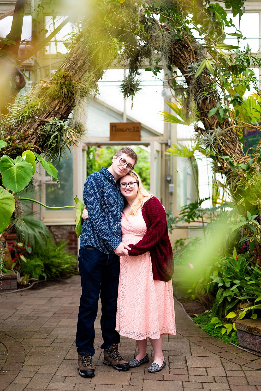 Couple posing together inside a lush greenhouse interior with brick flooring and tropical plants.