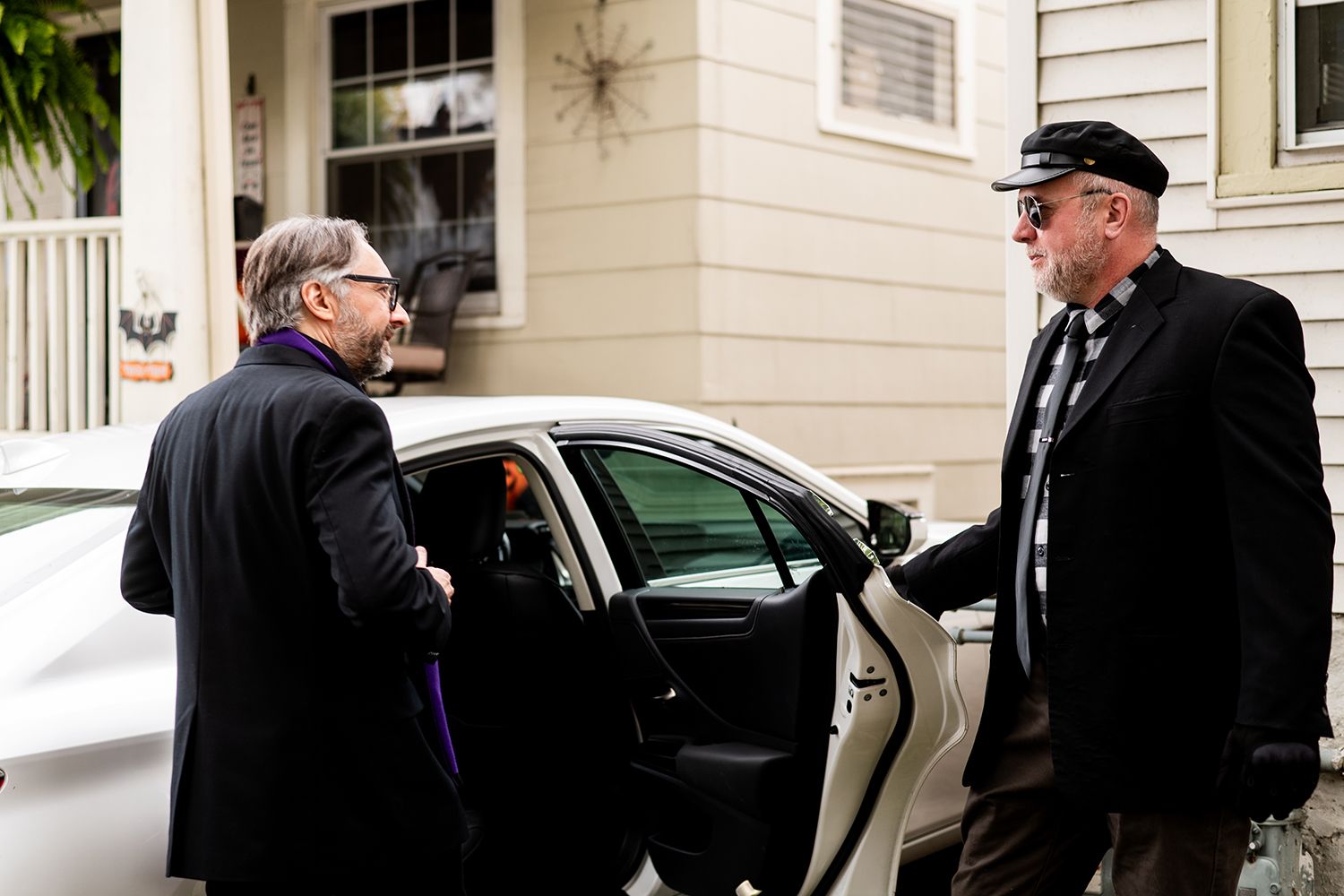 Several people in dark coats and sunglasses stand near white cars parked against a house.