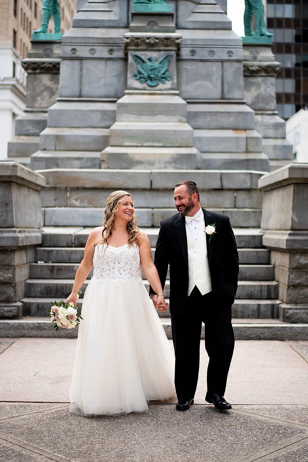 A joyful wedding couple walking together in front of stone steps with ornate architectural details.