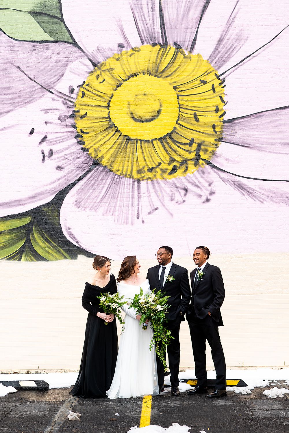 Wedding party stands beneath large floral mural of daisy with yellow center and white petals on urban wall.