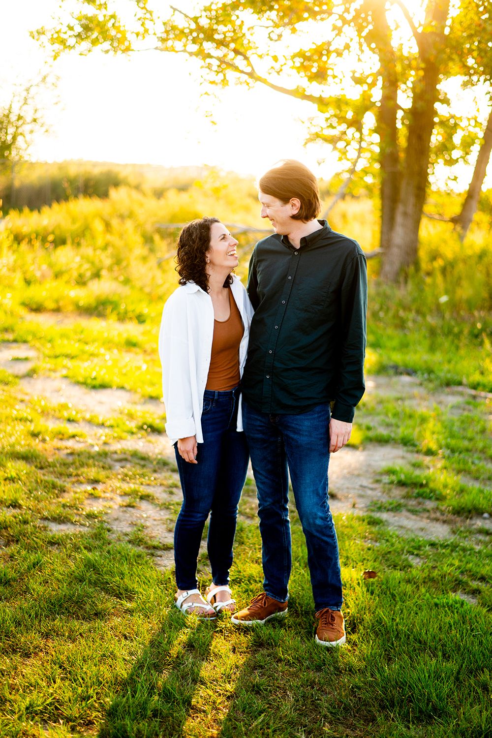 A couple stands together on a path during golden hour with sunlit trees and grass in the background.