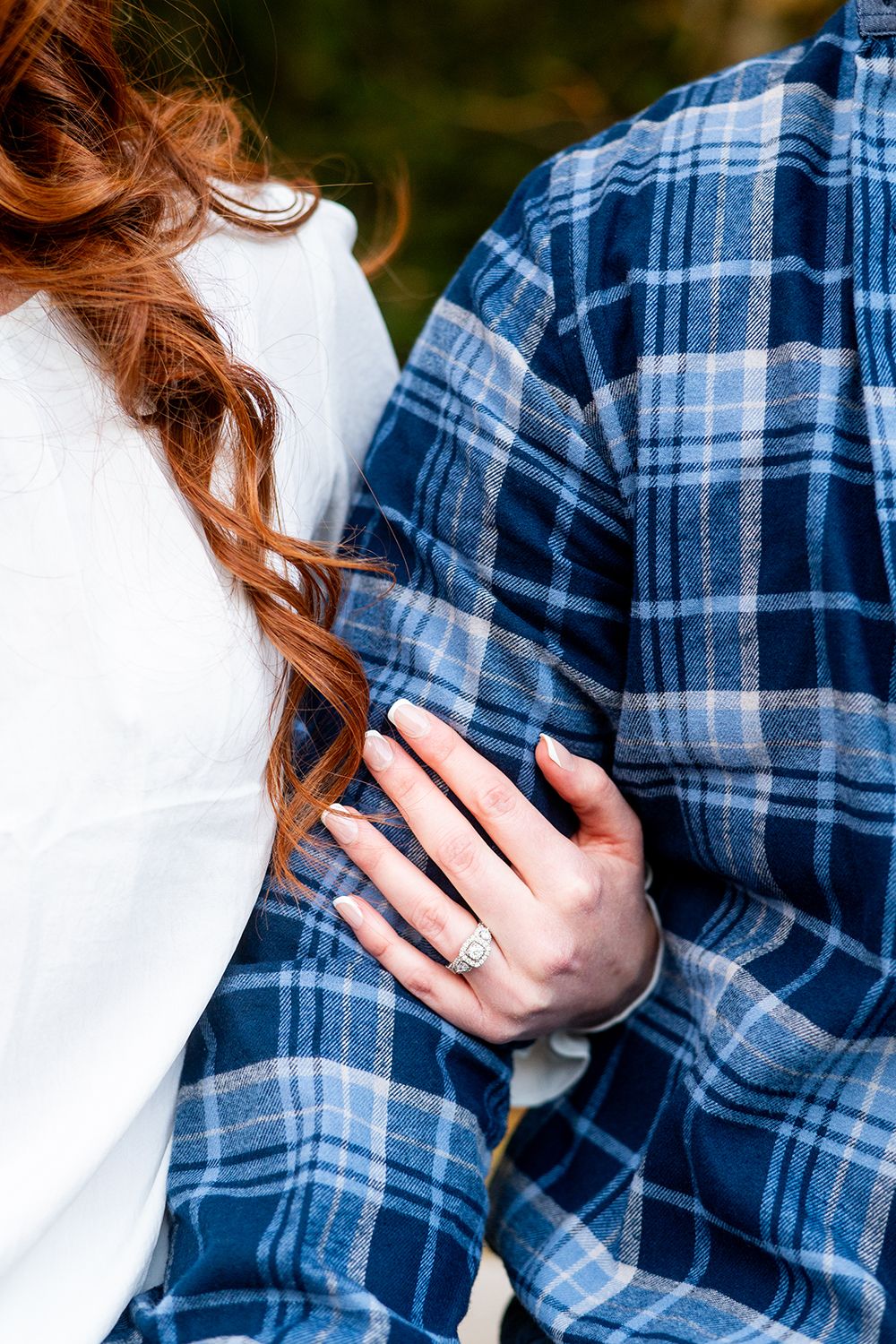 Person wearing a white top and blue plaid flannel shirt showing an engagement ring on their hand.