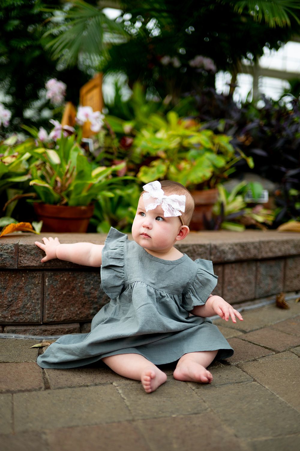 Baby in sage green dress sits on brick patio surrounded by lush garden foliage.