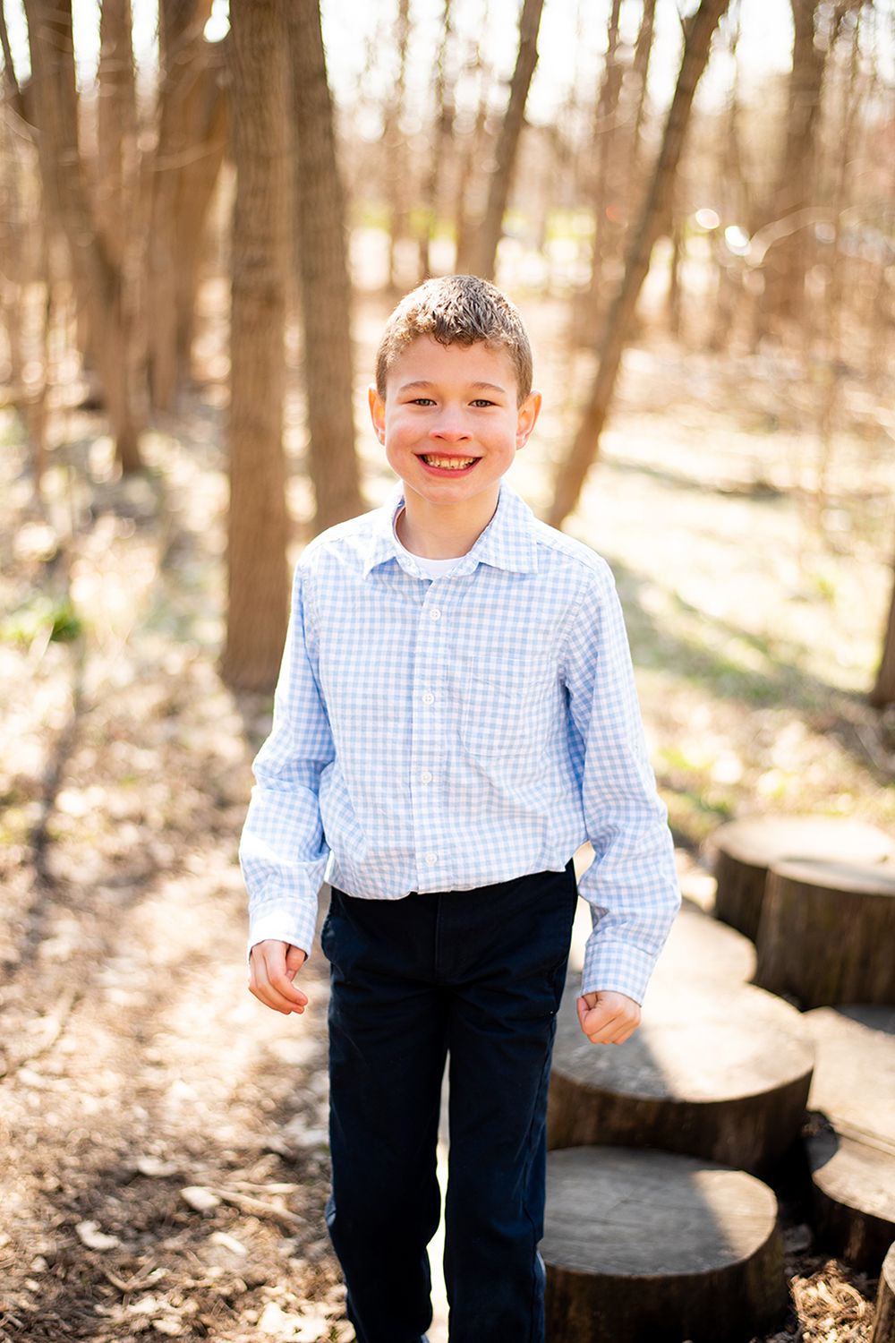 Young boy in blue checkered shirt standing on wooden steps in a sunny wooded area.