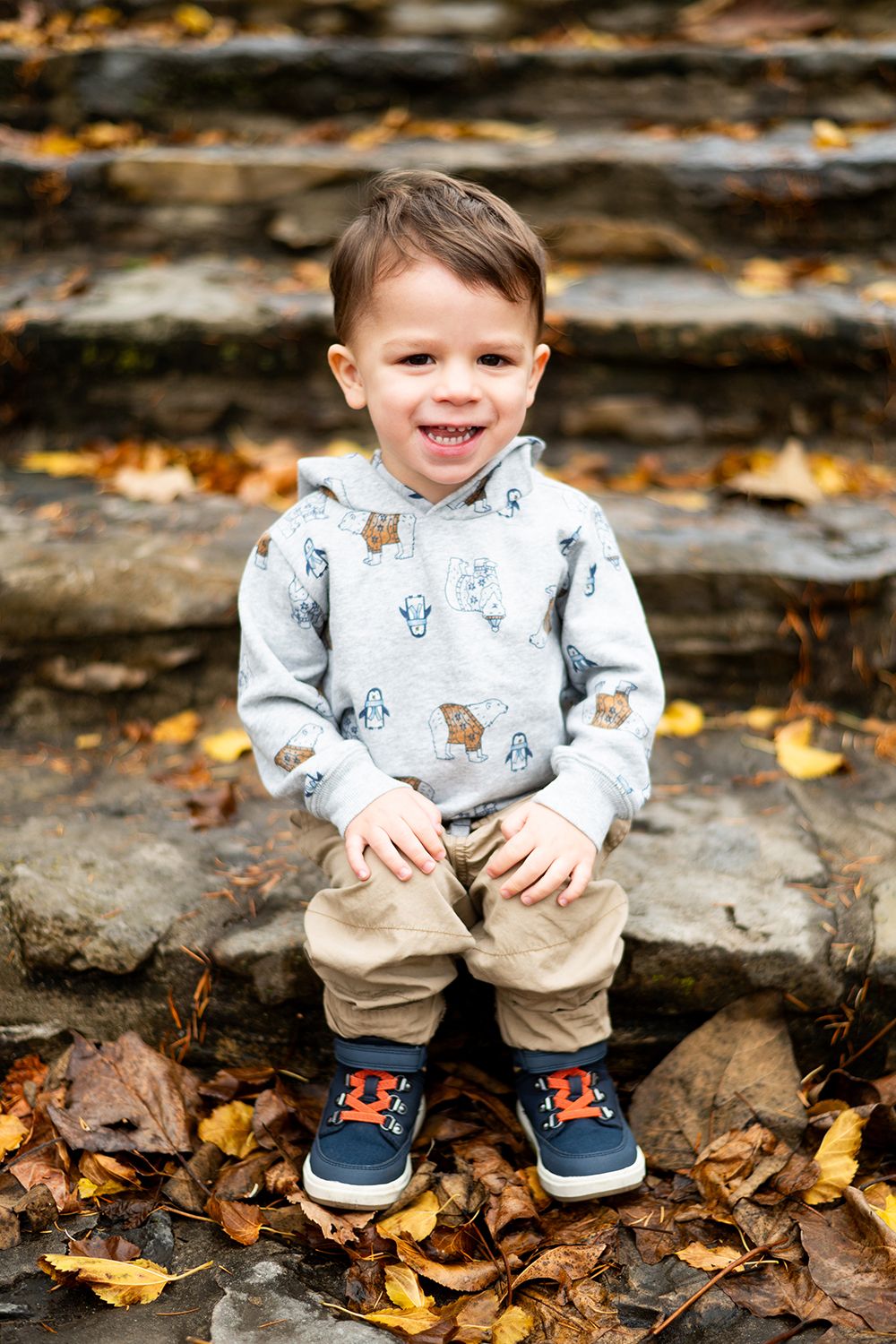 A toddler sitting on stone steps surrounded by autumn leaves wearing a printed sweatshirt and khaki pants.