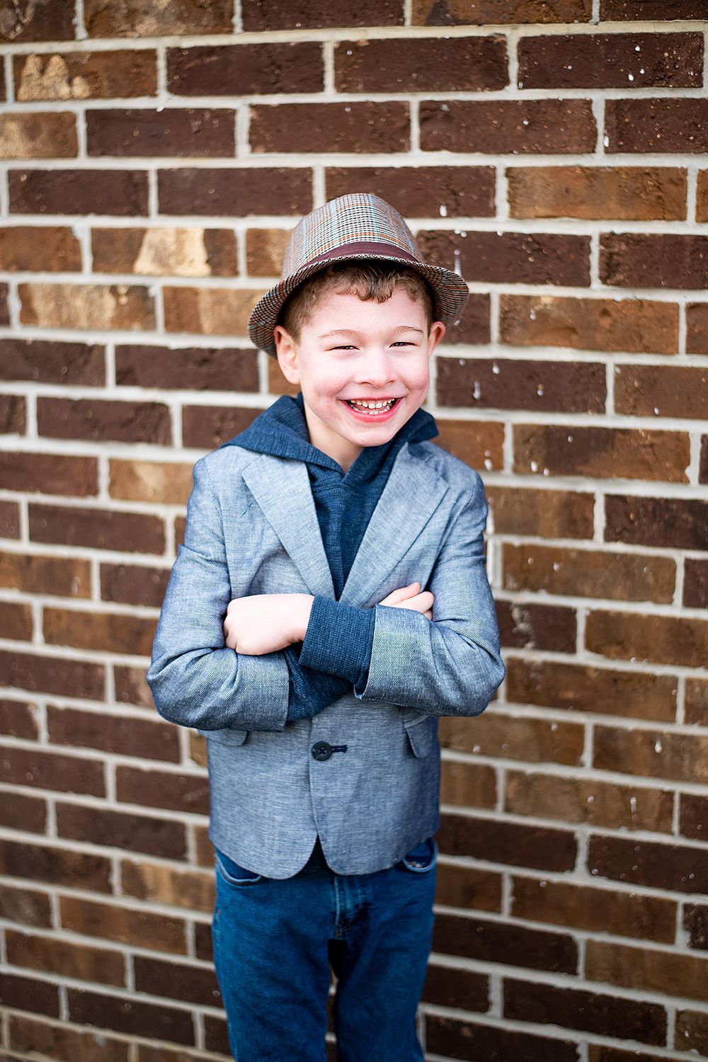 Young boy wearing a gray blazer, fedora hat and denim poses with crossed arms against brick wall.