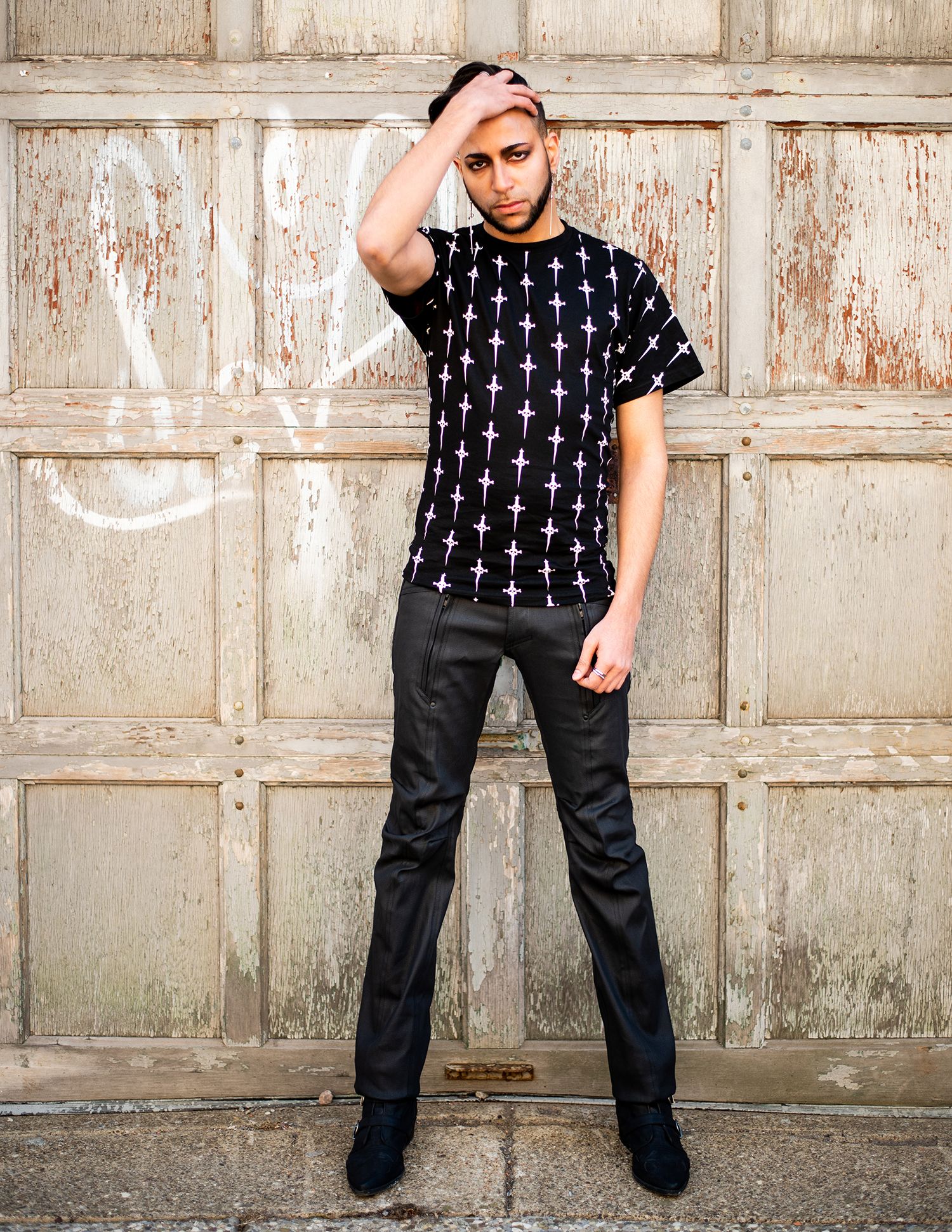 Someone wearing a black t-shirt with cross pattern and dark pants poses against a weathered wooden door.