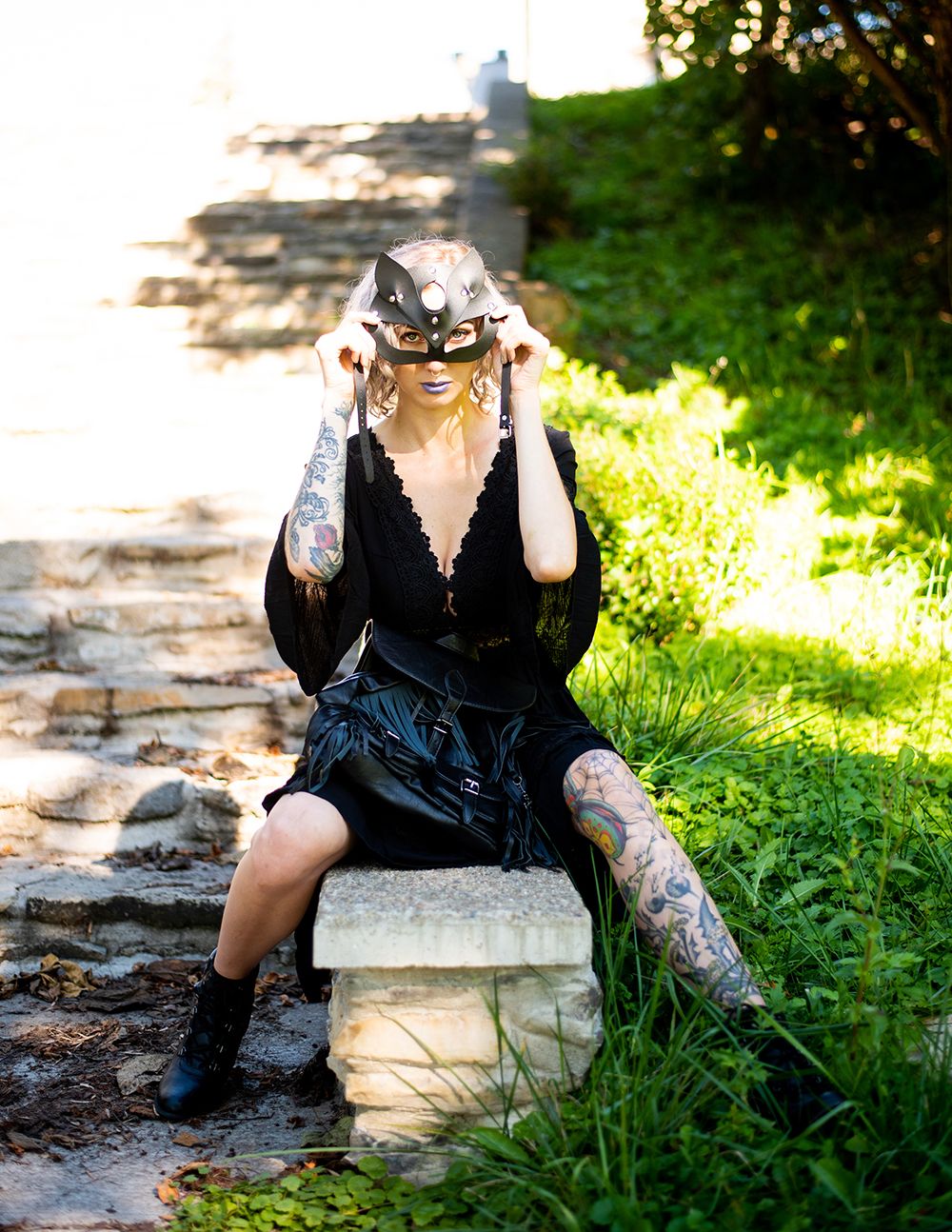 Person in a black outfit with tattoos sitting on stone steps in sunlit outdoor setting alongside a grassy path.