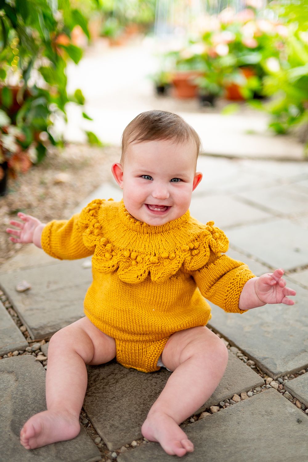 Baby in bright yellow knit sweater sits on stone pavers in sunny garden setting.