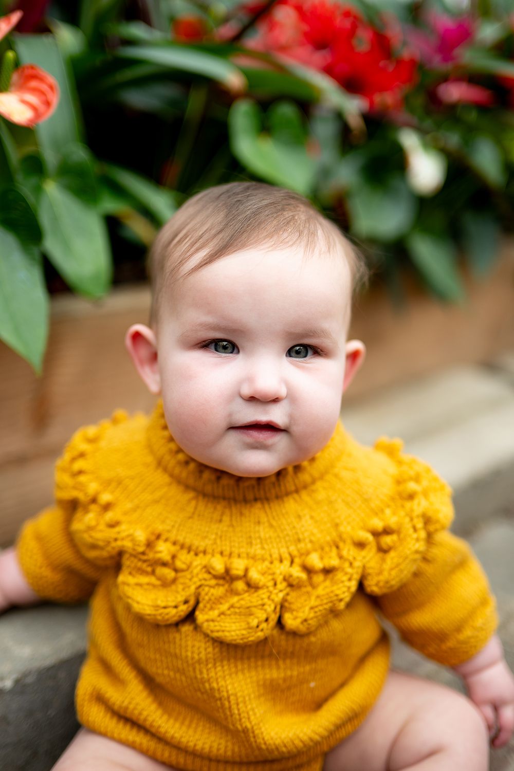 Close-up portrait of an infant in a bright yellow knit sweater with decorative ruffled neckline against floral backdrop.