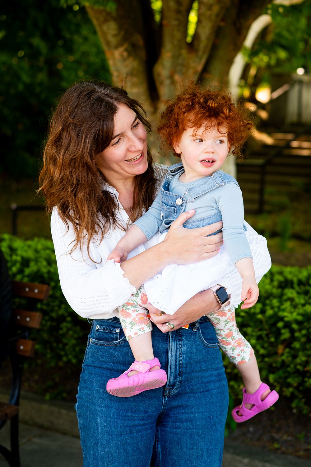 Woman in white shirt and jeans embraces a child wearing pink shoes in a garden setting.