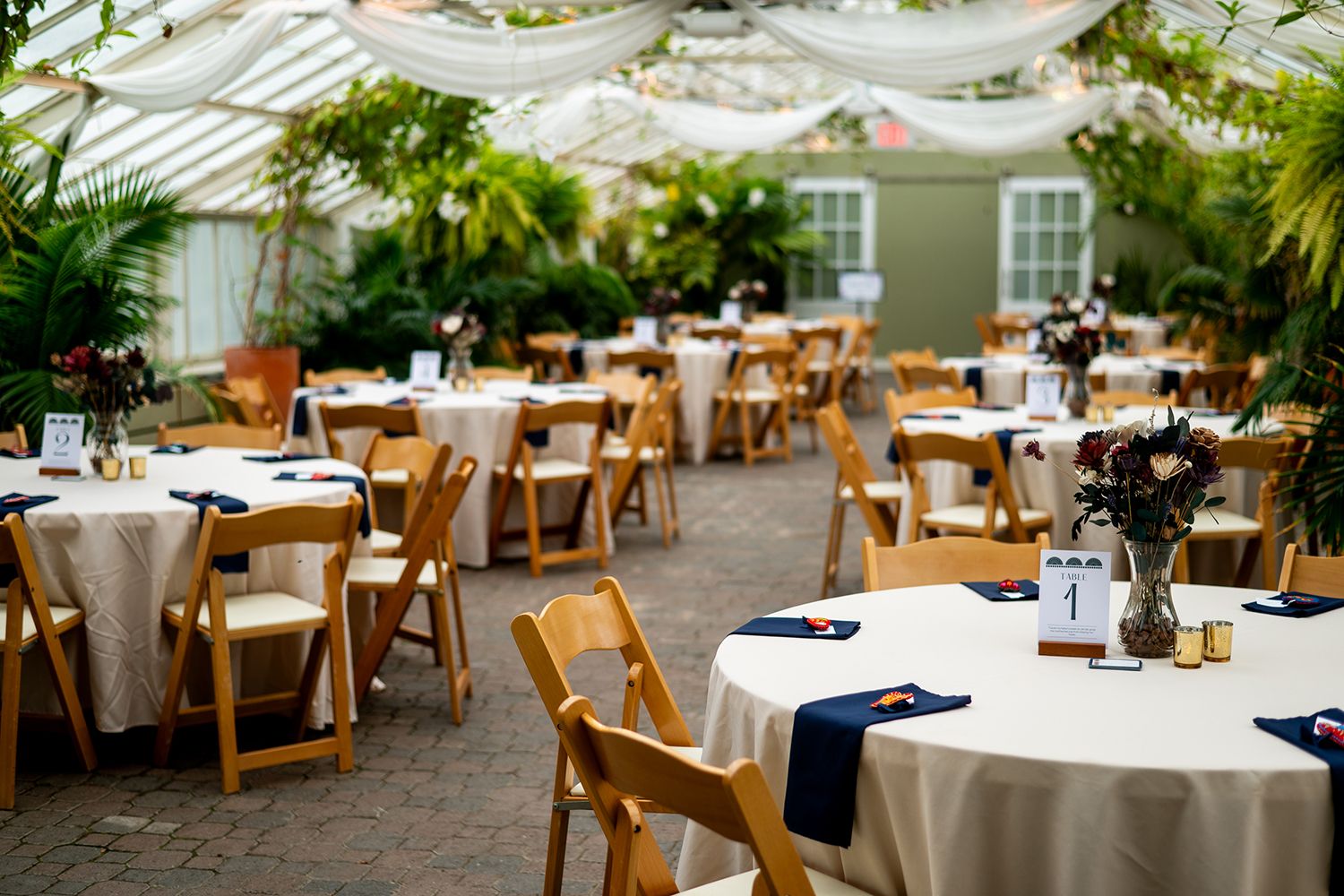 An elegant wedding reception setup with white-clothed tables and wooden chairs in a greenhouse-style venue.