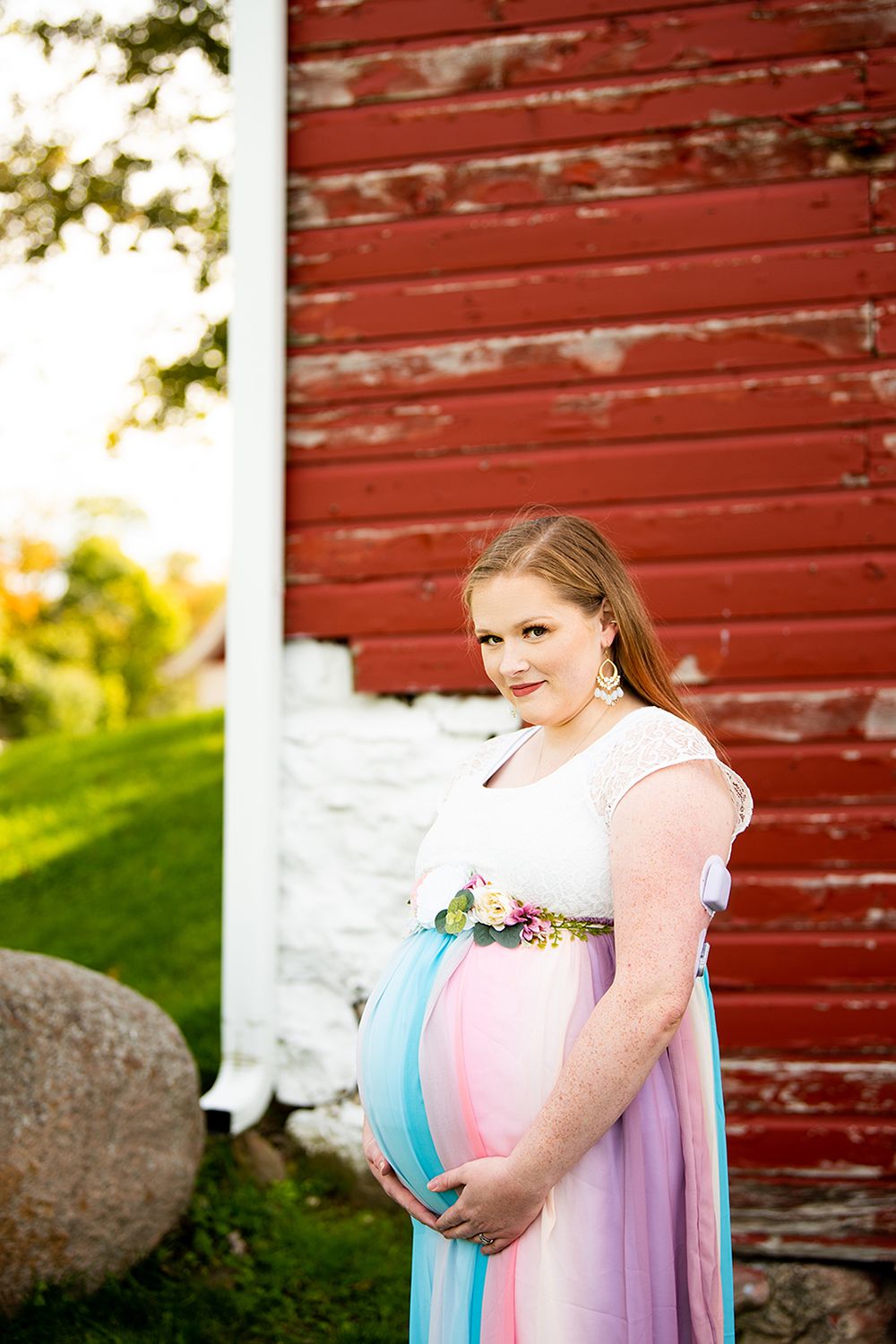 Maternity photos against a rustic red barn showing a woman in a pink and purple flowing dress during golden hour.
