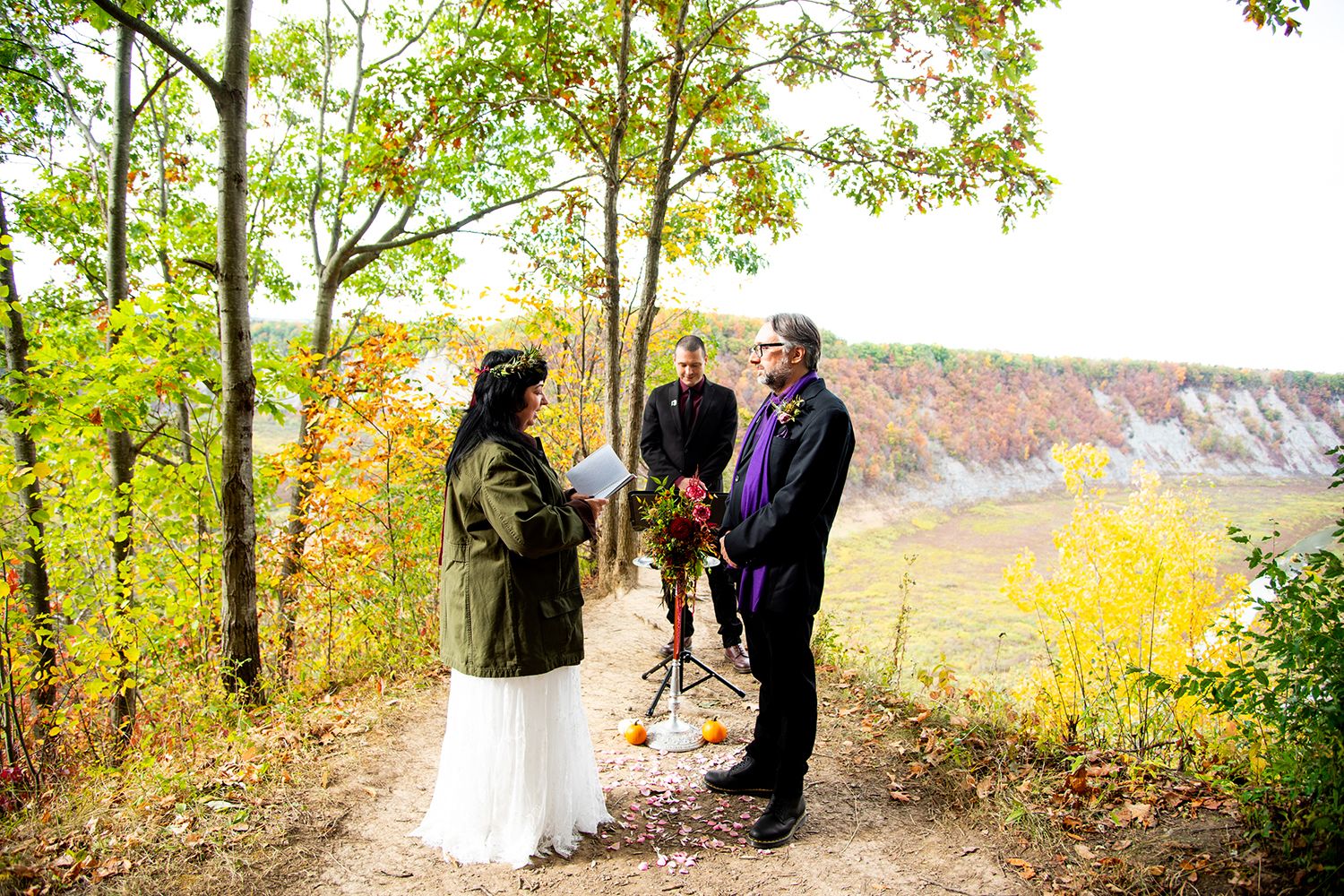 Wedding ceremony takes place on scenic wooded path with fall foliage in background.