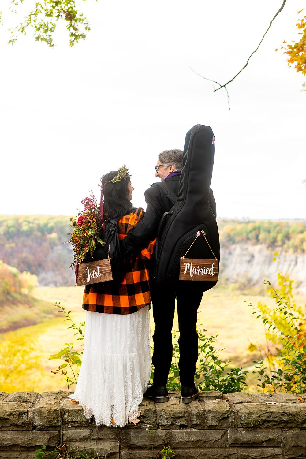 Couple in autumn wedding attire with guitar cases overlooking scenic fall valley from stone overlook.